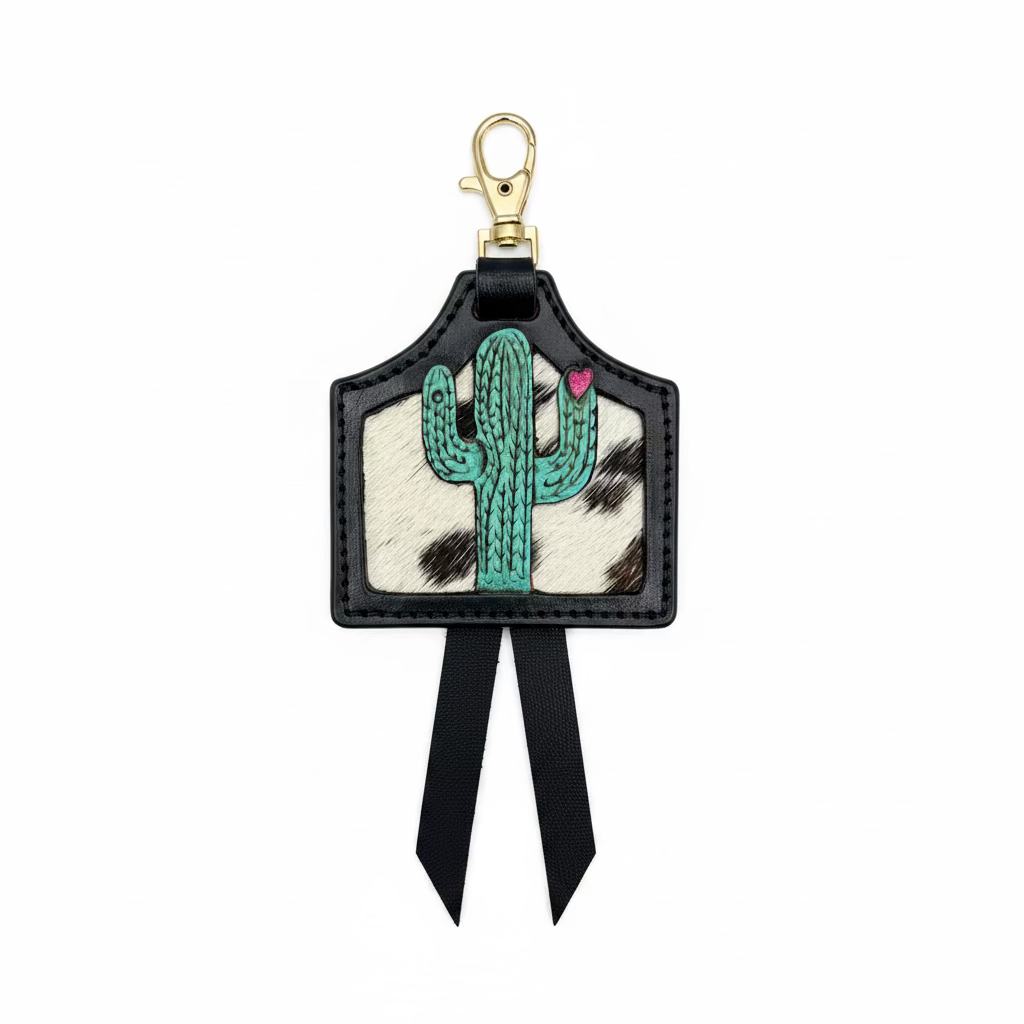 A western-style keychain or bag charm with a black leather border, a turquoise cactus on a white cowhide-patterned background, a small pink heart accent, and two black ribbon tails, displayed on a rustic wooden surface.