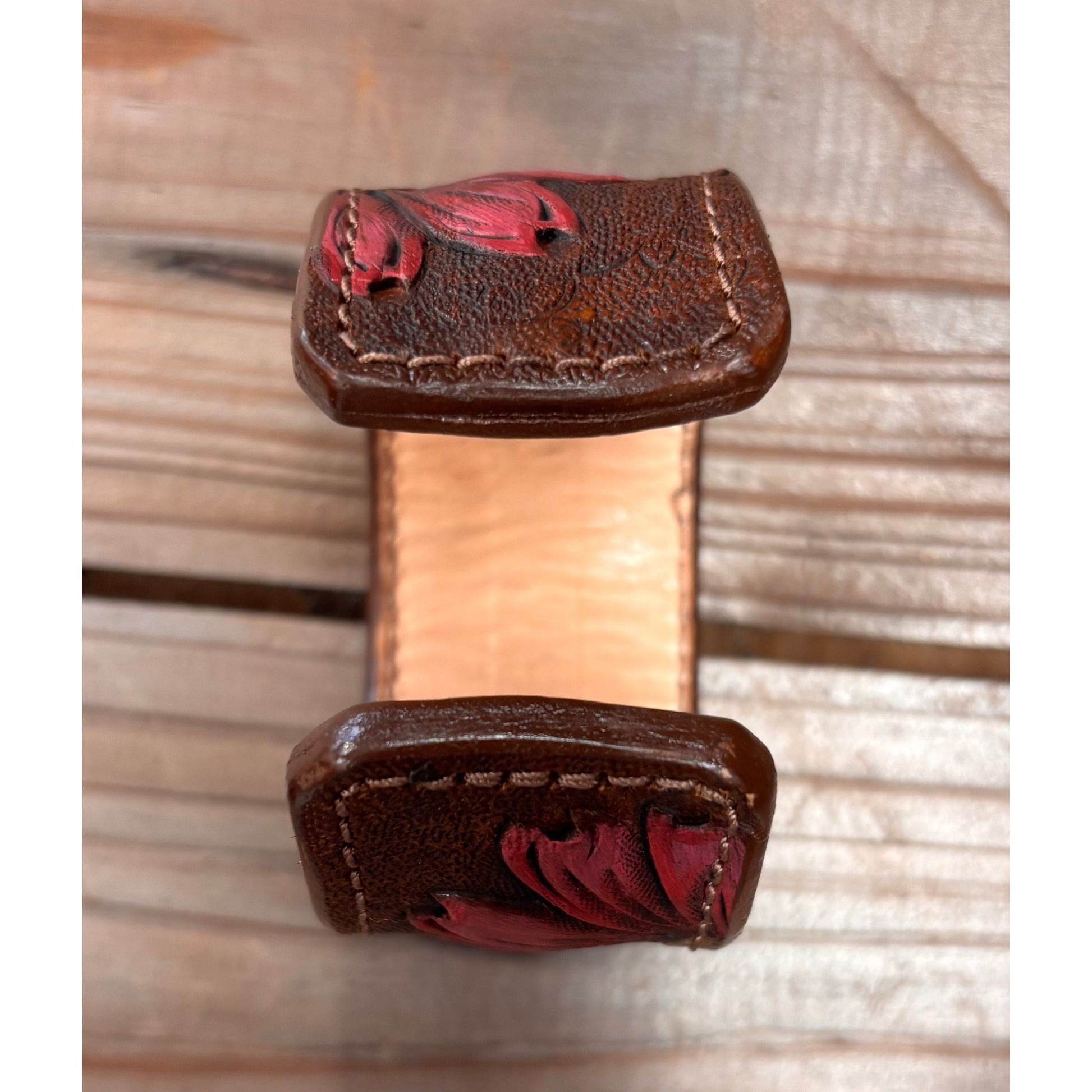 A close-up of a brown leather ring with red floral engravings on the flat sides, shown on a light wood background.