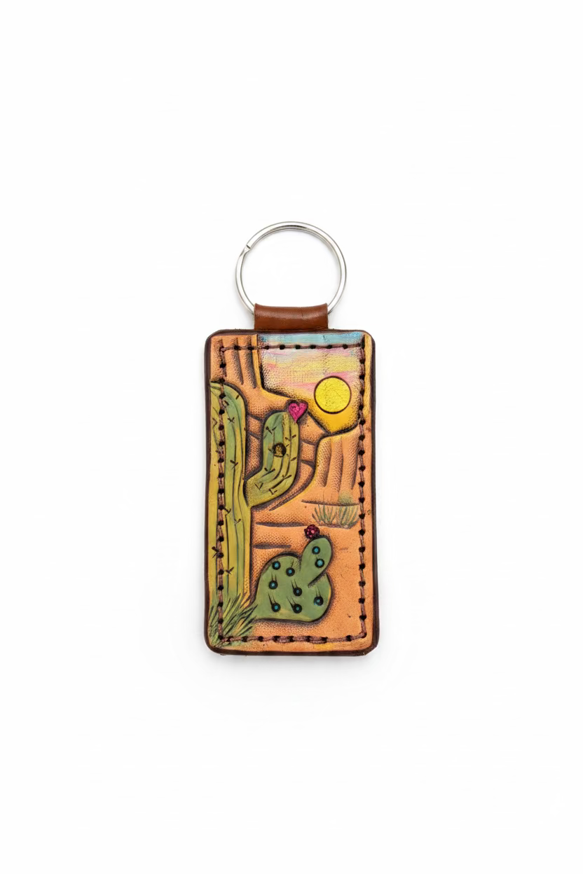 A rectangular hand-tooled leather keychain featuring a desert cactus design, resting on a rustic wooden surface.