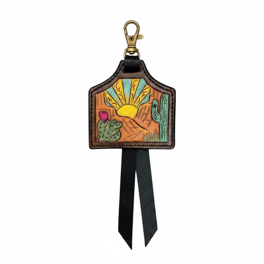 A hand-tooled leather keychain or bag charm with a desert landscape design, featuring a brass clasp and a black leather ribbon, displayed on a rustic wooden background.