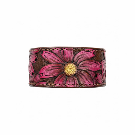 A hand-tooled leather cuff bracelet featuring a large pink and magenta daisy flower with a gold center, displayed on a rustic wooden surface.