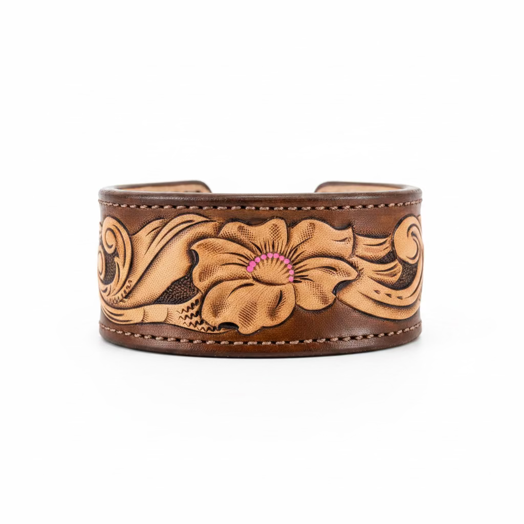 A wide brown leather cuff bracelet with hand-tooled floral designs and a small pink accent in the center, photographed on a rustic wooden surface.