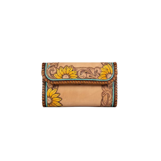 A hand-tooled leather clutch bag with a warm tan finish, featuring carved floral scrollwork and bright yellow sunflower designs, finished with turquoise-colored edging and braided brown leather trim, shown on a rustic wooden surface.