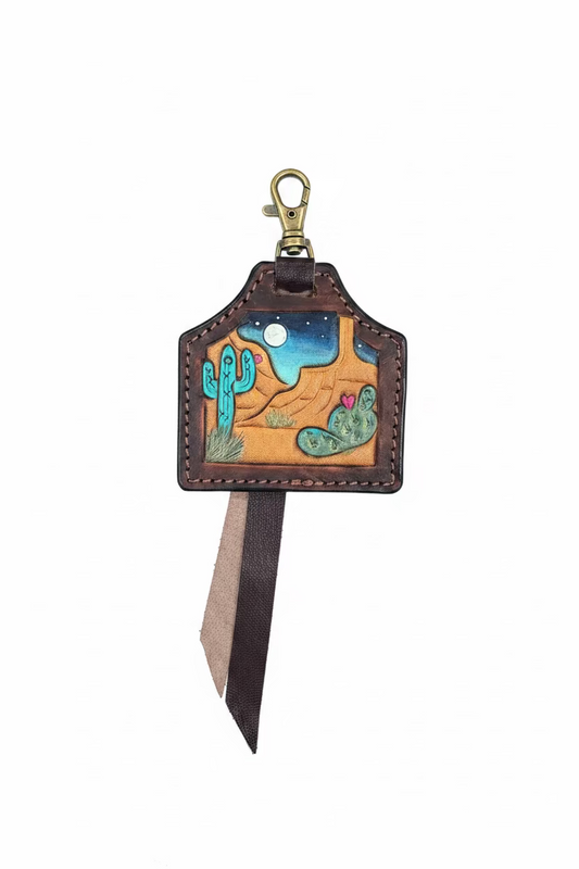 A handcrafted leather keychain or bag charm with a desert landscape design, featuring a brass clasp and a long leather ribbon tail, displayed on a dark gray background.