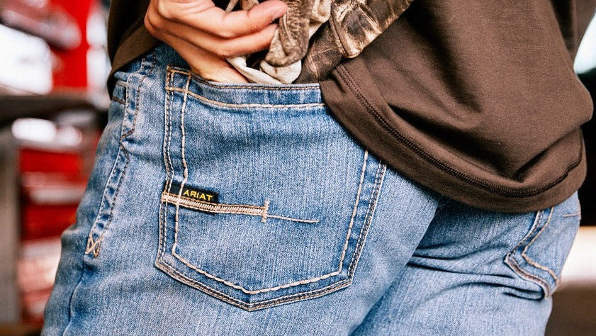 Person wearing blue jeans with a brand logo, holding a brown jacket.