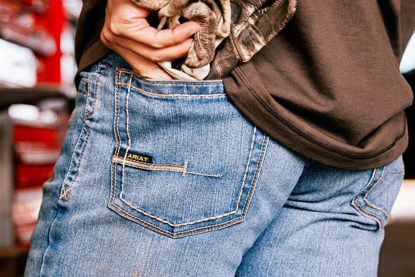Person wearing blue jeans with a brand logo, holding a brown jacket.